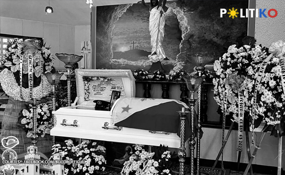 Black-and-white funeral scene with a flag-draped coffin surrounded by flowers and a religious altar featuring a cross and a central image of Jesus.