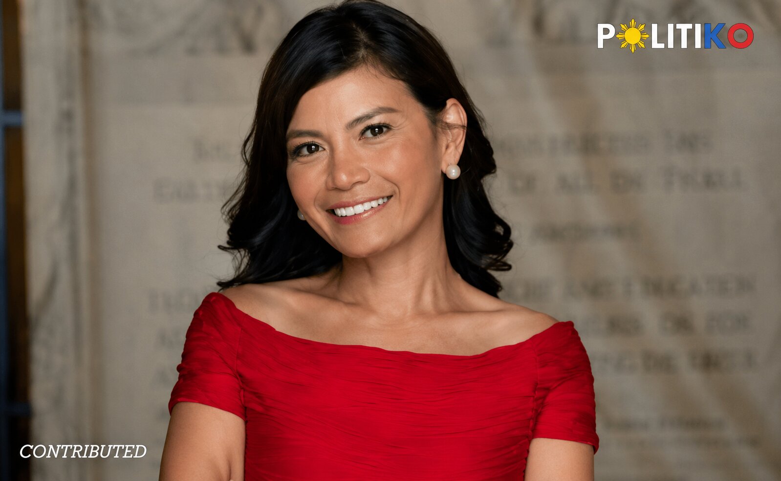 Smiling woman in a red off-the-shoulder dress poses for a portrait, with a POLITIKO watermark in the top-right corner.