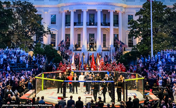 A large fenced mixed martial arts cage set up on the White House lawn, surrounded by a crowd and flag bearers, with the White House facade in the background and evening lighting.